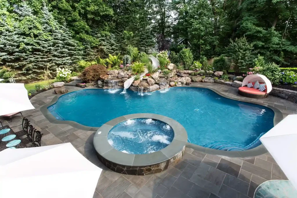 Freeform pool with raised circular spa, stone paver patio, rock waterfall, and slide surrounded by lush landscaping in New Jersey by Gorlin Pools