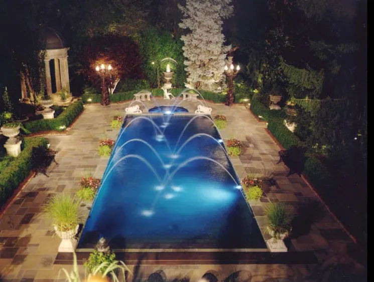 Aerial night view of a rectangular pool with arc water jet fountains, underwater lighting, and formal garden statuary in Lakehurst, NJ by Gorlin Pools