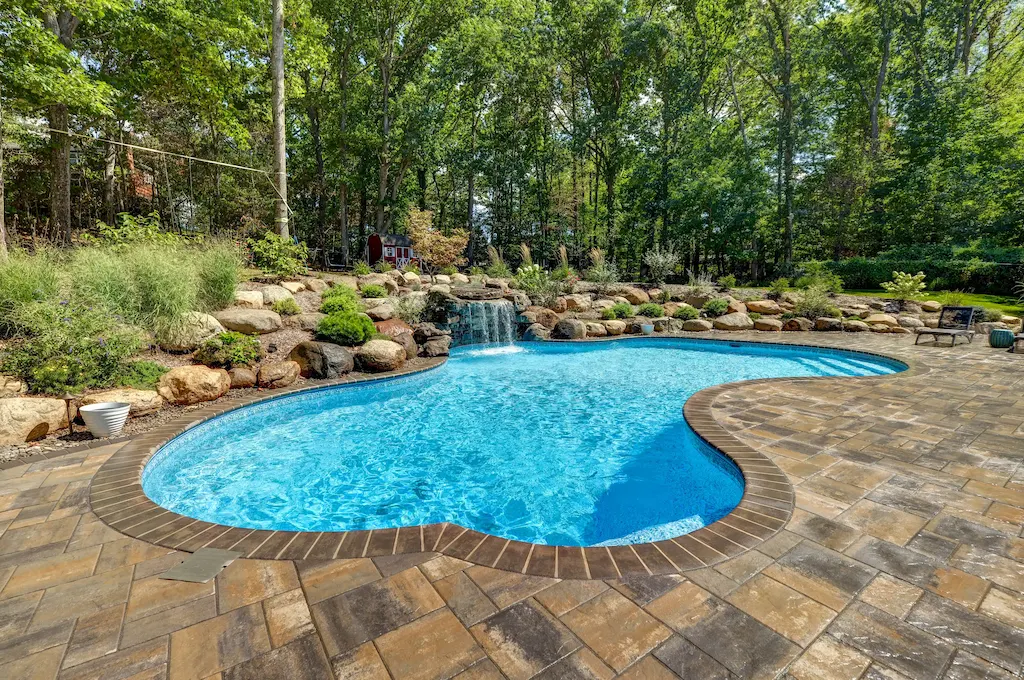 Freeform pool with natural rock waterfall, boulder landscaping, and paver deck in wooded backyard in Lakehurst, NJ by Gorlin Pools