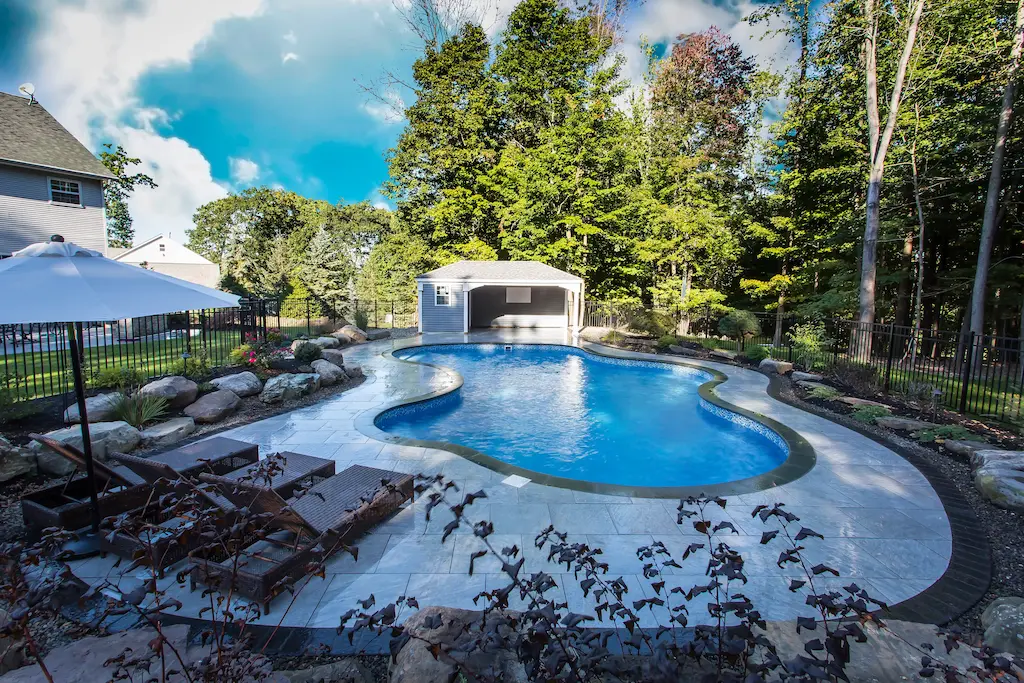 Lagoon pool with large tanning ledge, bluestone deck, and gray pool house cabana with boulders in Lakehurst, NJ by Gorlin Pools