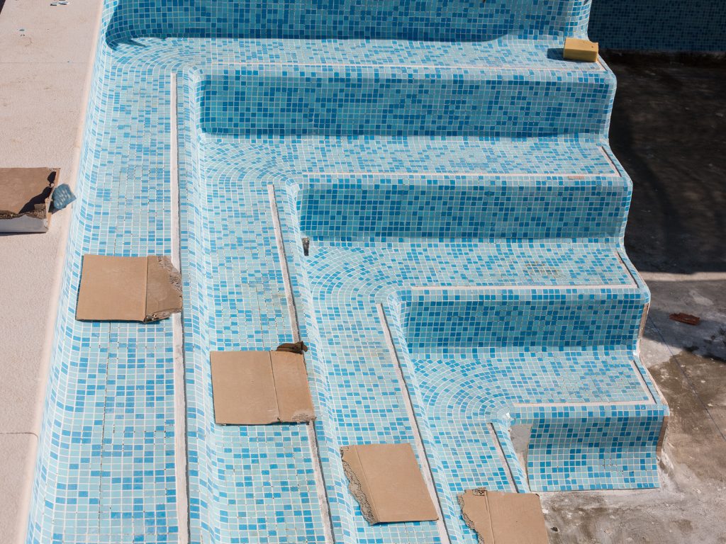 Steps of an empty swimming pool feature blue mosaic tiles. Cardboard squares cover some steps, indicating ongoing repairs, with a sunny, calm atmosphere.