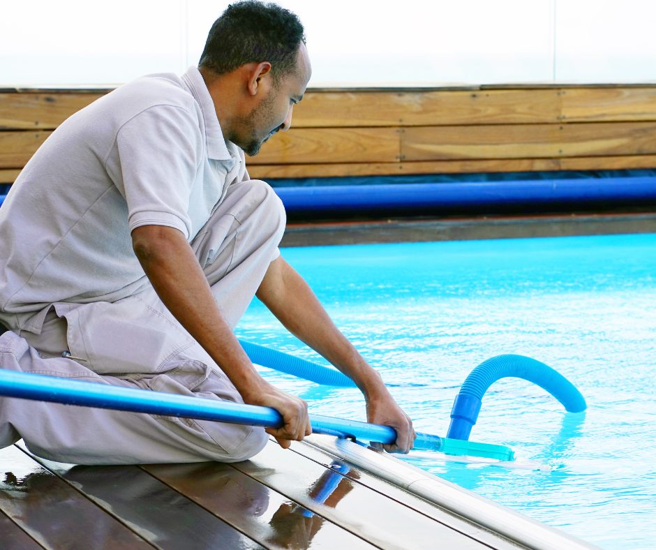 A man in a light uniform kneels on a wooden deck, cleaning a clear blue pool with a long pole and vacuum hose, focused and intent on his work.