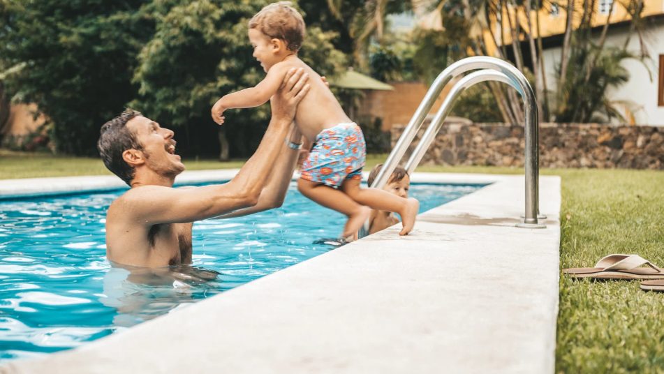 A joyful man in a pool lifts a smiling toddler, who is wearing colorful swim trunks, at the pool edge. Bright, sunny ambiance with green grass nearby.