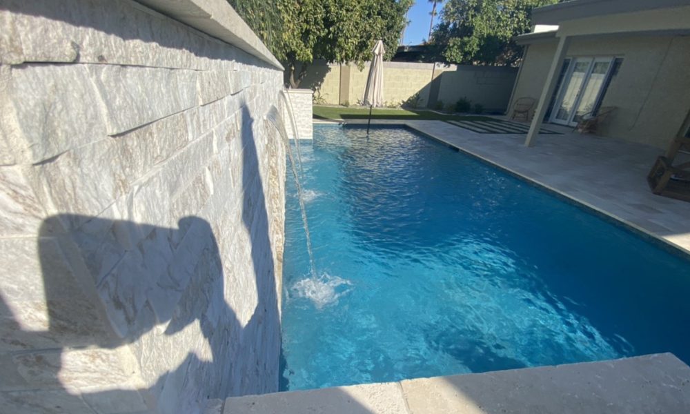 A serene backyard pool with a stone wall and water fountain feature. Sunlight casts shadows on the wall, creating a calm and inviting atmosphere.