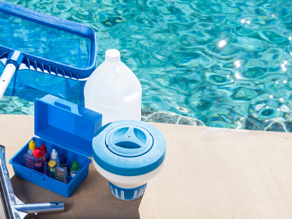 Pool maintenance tools on a sunny day: a skimmer net, open test kit, large white bottle, and floating chlorine dispenser beside a clear blue pool.