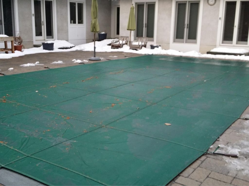 A snow-dusted backyard patio with a large green pool cover, surrounded by patio chairs and closed umbrellas. The scene conveys a chilly winter atmosphere.