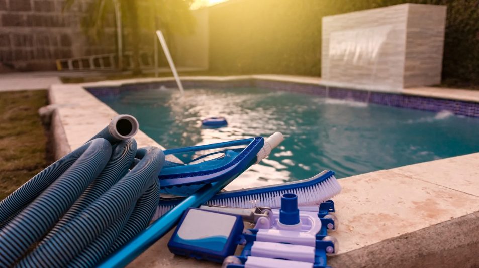 Pool cleaning tools including hoses, a brush, and a vacuum attachment are placed on the edge of a blue-tiled pool. The scene is warmly lit by the setting sun.