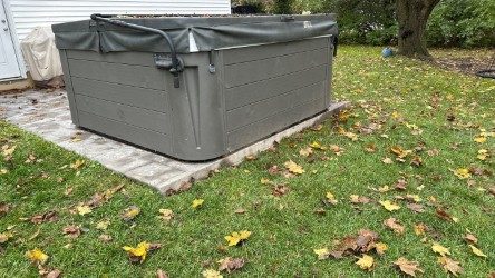 Covered outdoor hot tub on a patio in the backyard. Surrounded by grass and scattered yellow leaves, suggesting an autumn setting. Calm, serene atmosphere.