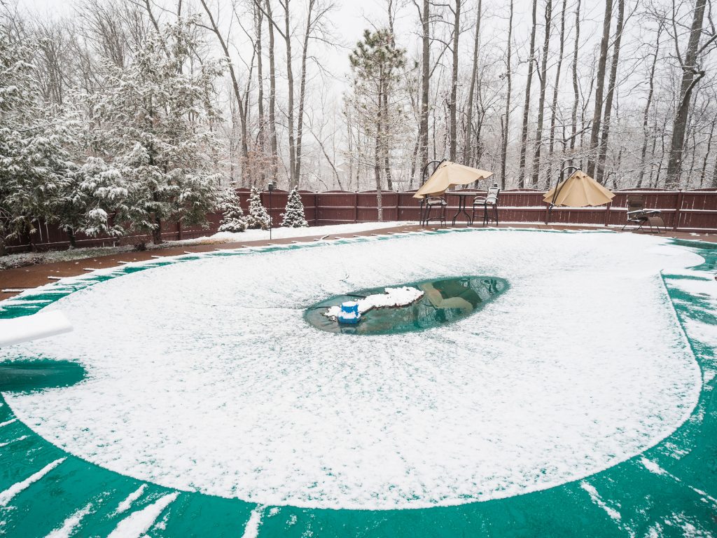 Snow-covered pool with a green cover surrounded by a snowy yard and trees. Patio furniture with umbrellas sits near a red wooden fence, creating a serene winter scene.