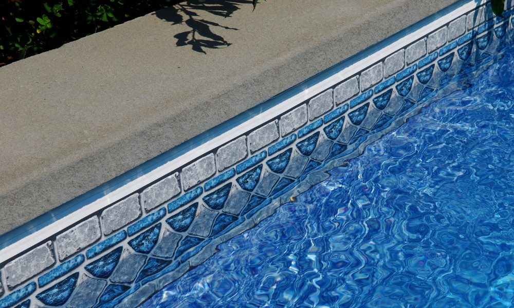 Clear blue swimming pool water with sunlight reflections, bordered by a decorative tile pattern under a concrete edge. Shadow of leaves on the concrete.
