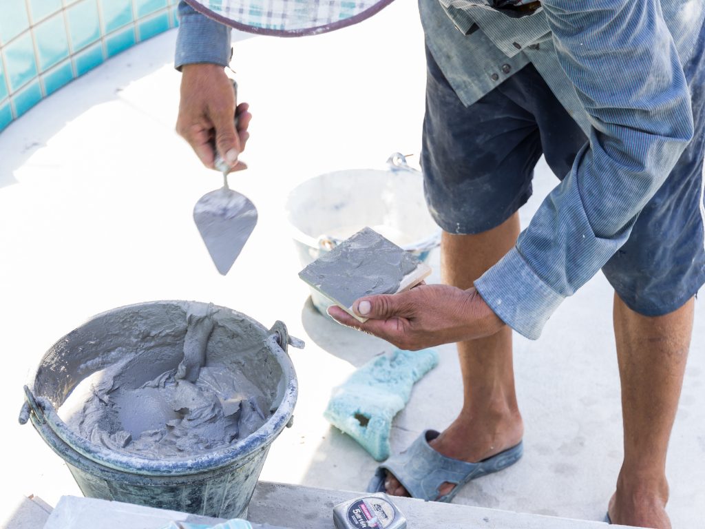 A worker wearing blue clothes is applying gray mortar to a tile using a trowel. A bucket of mortar, sponge, and measuring tape are nearby on the ground.
