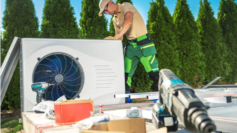 A technician in a beige shirt and green pants installs an outdoor air conditioning unit. Tools are on a surface nearby, with trees in the background.