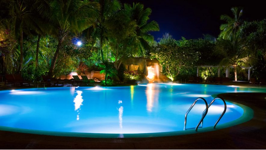A tranquil pool at night, surrounded by lush, illuminated palm trees and foliage. The water glows vibrant blue, conveying a serene and inviting atmosphere.