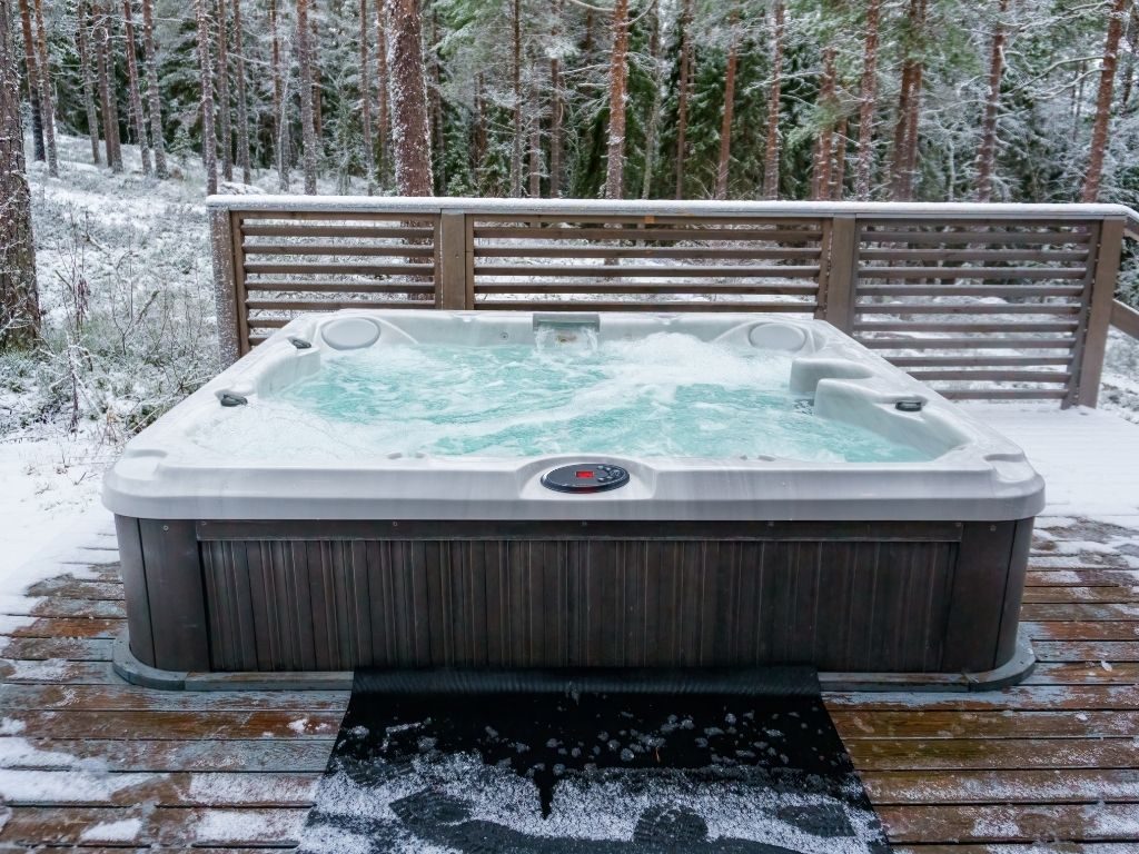 Outdoor hot tub bubbling on a snowy wooden deck, surrounded by a wintry forest. The scene conveys a cozy, contrasting warmth and cold.